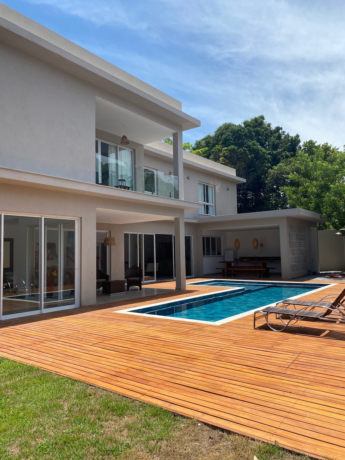 The two-story house features a modern exterior with large glass doors. A wooden deck surrounds the inviting pool, while lush greenery adds a natural backdrop. Ample seating is arranged nearby, providing an ideal space for relaxation.