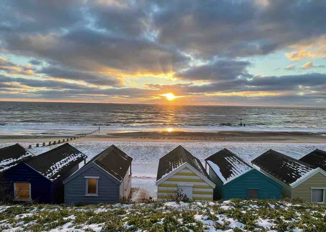 The image depicts a winter beach scene at sunset, with a row of colorful beach huts in the foreground. The snow-covered sand and sea reflect the warm hues of the setting sun, while clouds gather in the sky above, creating a serene coastal atmosphere.