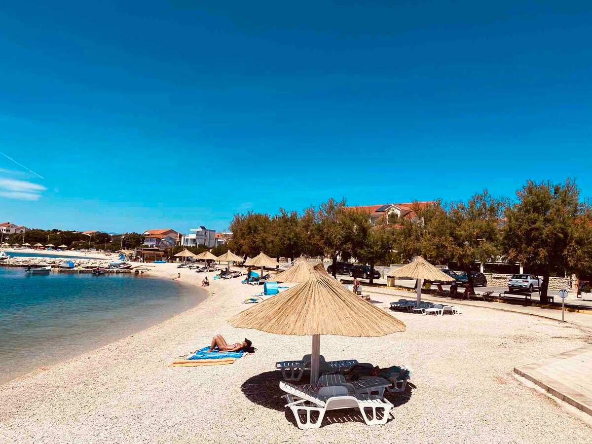 A sandy beach extends along the shoreline with straw umbrellas providing shade. Sun loungers are positioned under the umbrellas. The clear blue sky overhead complements the serene atmosphere, while boats are anchored in the calm waters, and a distant row of trees lines the beach.