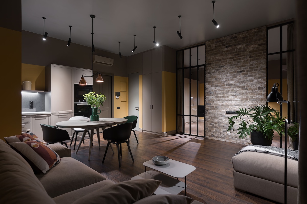A contemporary living area features a cozy sofa with colorful cushions, a sleek dining table surrounded by chairs, and stylish lighting overhead. A brick wall adds texture to the space, while potted plants bring a touch of greenery. The kitchen area is subtly integrated in the background.