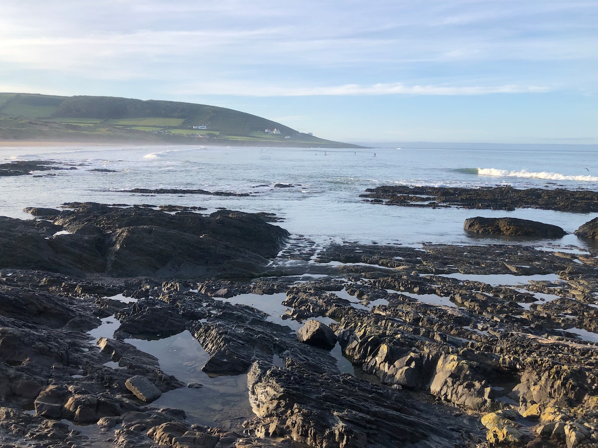 The rocky shoreline is displayed with smooth wet stones and tidal pools reflecting the sky. Gentle waves are seen lapping against the rocks, while a distant hillside with greenery provides a serene backdrop. Clear skies and soft lighting contribute to the tranquil scene.
