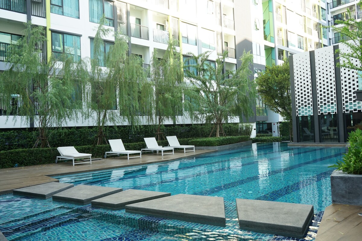 A serene swimming pool is featured, characterized by clear blue water and surrounded by elegant lounge chairs. Stone stepping platforms create a pathway across the pool, while lush greenery and residential buildings in the background enhance the tranquil ambiance.