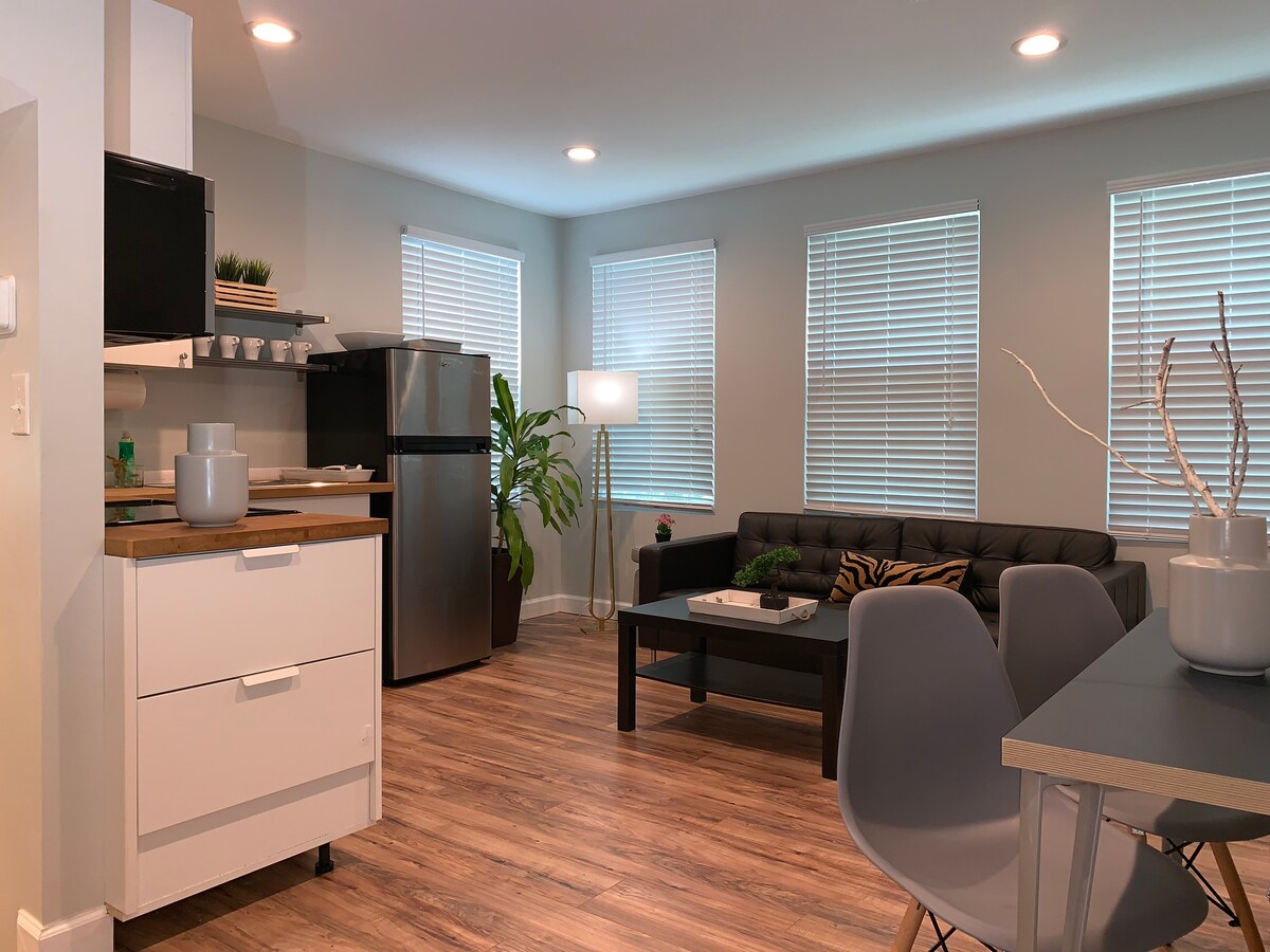 The spacious living area features light grey walls and modern furnishings. A dark leather couch is positioned near large windows with blinds, complemented by a potted plant. A wooden countertop separates the kitchen with a stainless steel refrigerator, and a dining table with simple grey chairs is nearby.