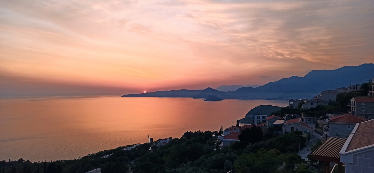 A panoramic view of a coastal landscape is displayed at sunset, with soft orange and pink hues reflected on the calm water. Silhouettes of distant mountains and coastal homes are visible, contributing to a serene and tranquil atmosphere.