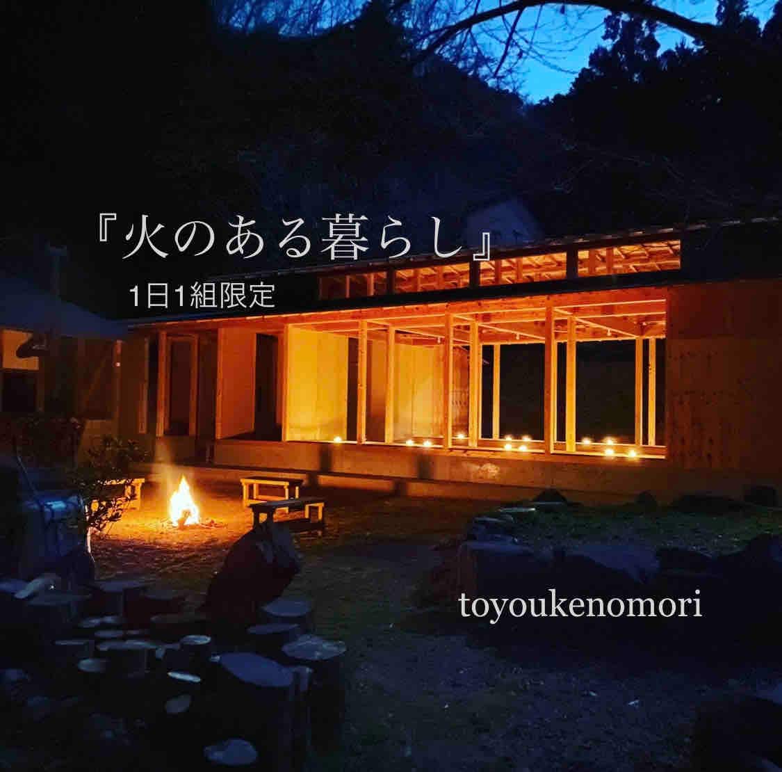 A serene outdoor scene is captured at dusk, featuring a cozy fire pit surrounded by simple seating. Soft candlelight illuminates a traditional wooden structure, creating a calming ambiance against the backdrop of nature.