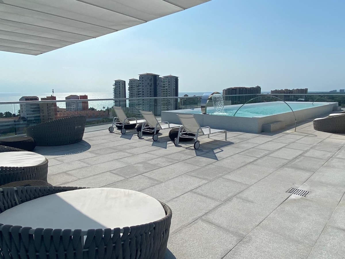 A rooftop terrace features a spacious area with lounge chairs arranged around a sparkling swimming pool. Comfortable circular seating is visible, against a backdrop of modern buildings and a clear blue sky. The design allows for relaxation and enjoyment of the stunning views.