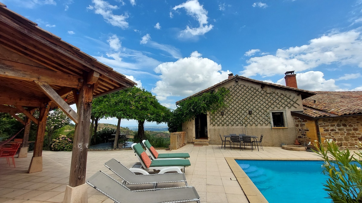 A serene outdoor area is visible, featuring a rectangular pool surrounded by several lounge chairs. Nearby, an inviting dining space is sheltered under a wooden structure, with a table and chairs set up for meals. Lush greenery and a calm sky add to the peaceful ambiance.