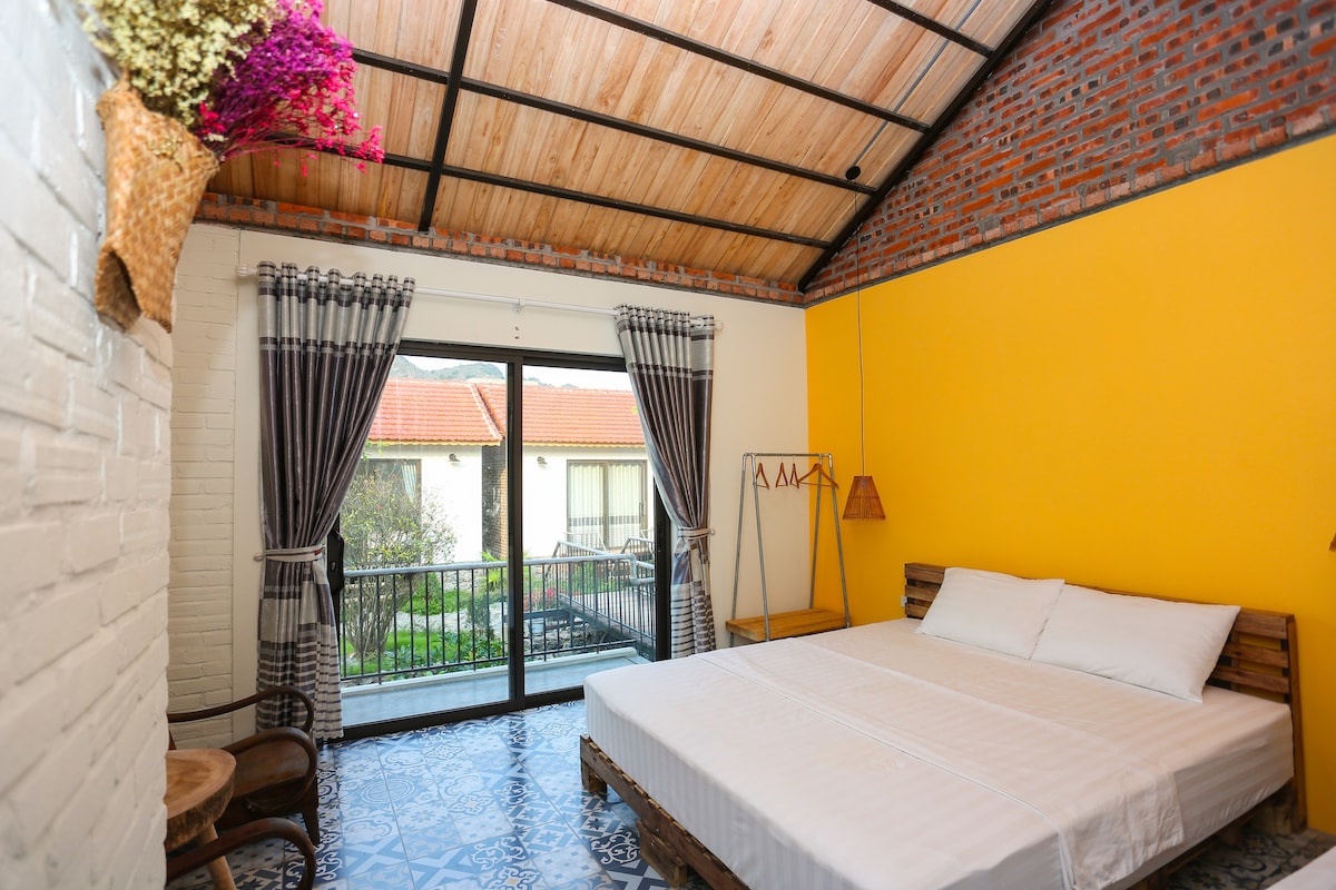 A brightly colored room features a spacious bed with white linens. Large glass doors provide access to a balcony, allowing natural light to fill the space. The walls are painted a lively yellow, complemented by decorative lighting and floral arrangements.