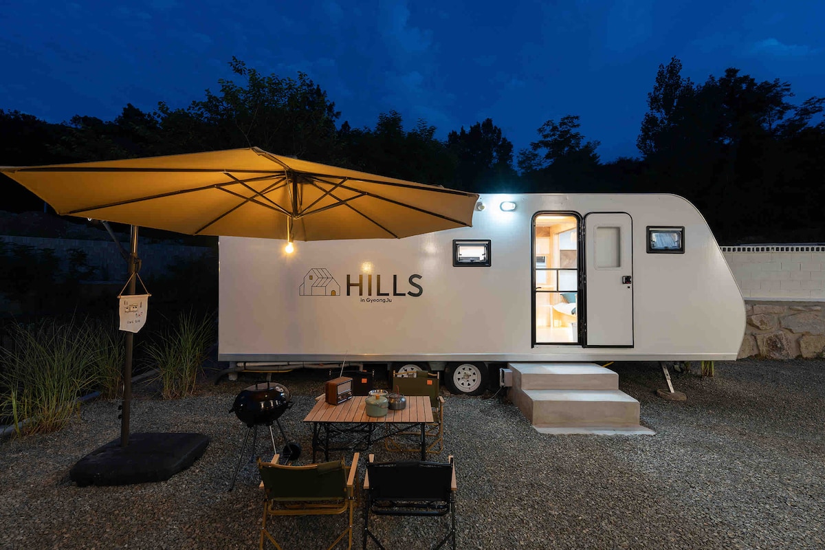 A caravan is positioned under a darkening sky, enhanced by an orange canopy providing shade over a small outdoor seating area. A table and chairs are set nearby, while the caravan features a welcoming entrance illuminated by soft lighting.
