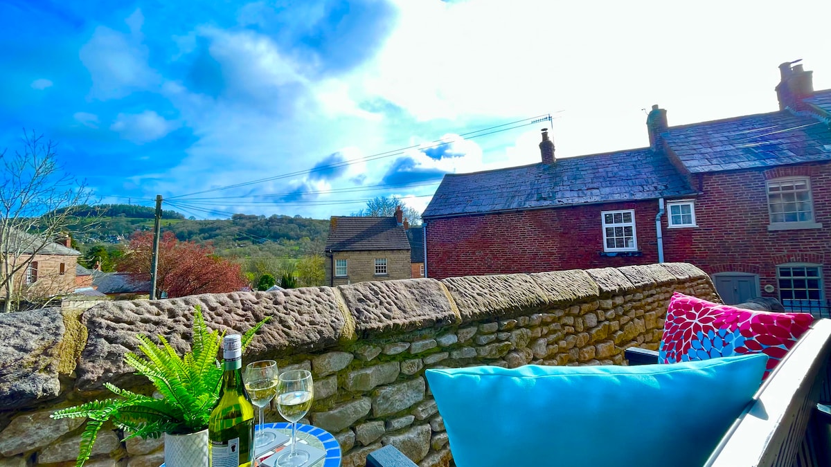 A view from a balcony showcases a stone wall and nearby brick houses, framed by rolling hills. Two glasses sit on a small table, accompanied by a green plant and colorful cushions, inviting relaxation in the fresh air.