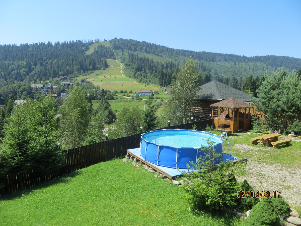 An outdoor area featuring a round blue pool set on a grassy lawn, surrounded by trees. A wooden gazebo to the right offers shaded seating, while a picnic table rests nearby. The background showcases a scenic view of rolling hills and forested mountains under a clear blue sky.