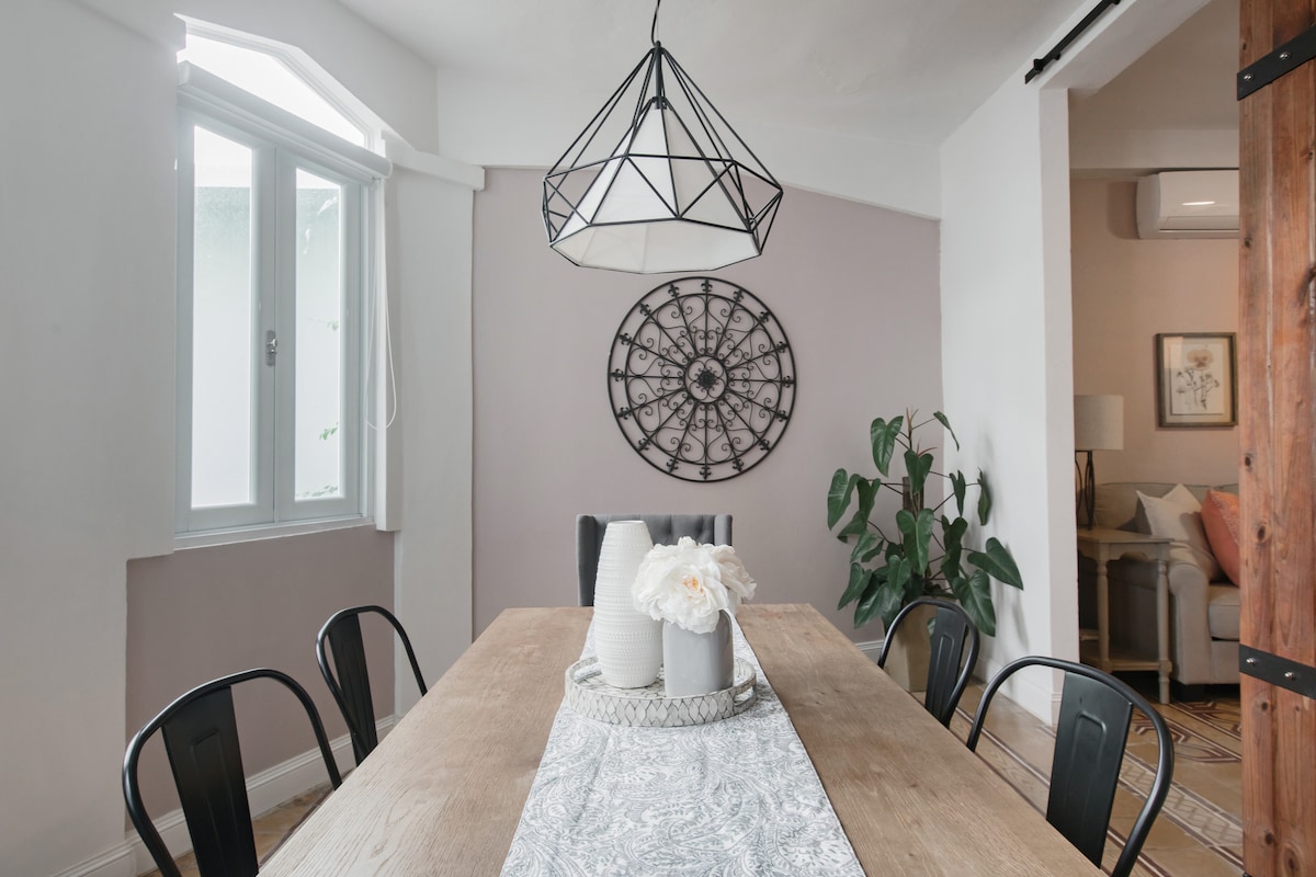 A dining area is presented with a large wooden table surrounded by black chairs. A geometric light fixture hangs above the table, which is adorned with a decorative runner and a centerpiece featuring vases of flowers. A large wall clock is displayed on the pale wall, enhancing the space.