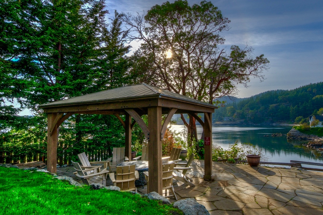 A shaded gazebo offers seating surrounded by natural stone, overlooking calm waters. Lush greenery frames the scene, with sun rays creating a serene ambiance. The stone pathway leads to the water's edge, inviting relaxation in a tranquil outdoor setting.