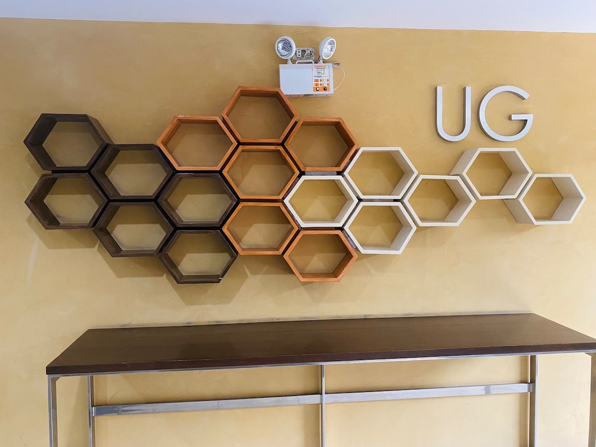 A wall-mounted honeycomb shelf design features various shades of wood, including brown, orange, and cream. The arrangement creates a modern visual contrast against the beige wall. Above the shelf, the letters 'UG' are prominently displayed, adding to the contemporary aesthetics of the space.