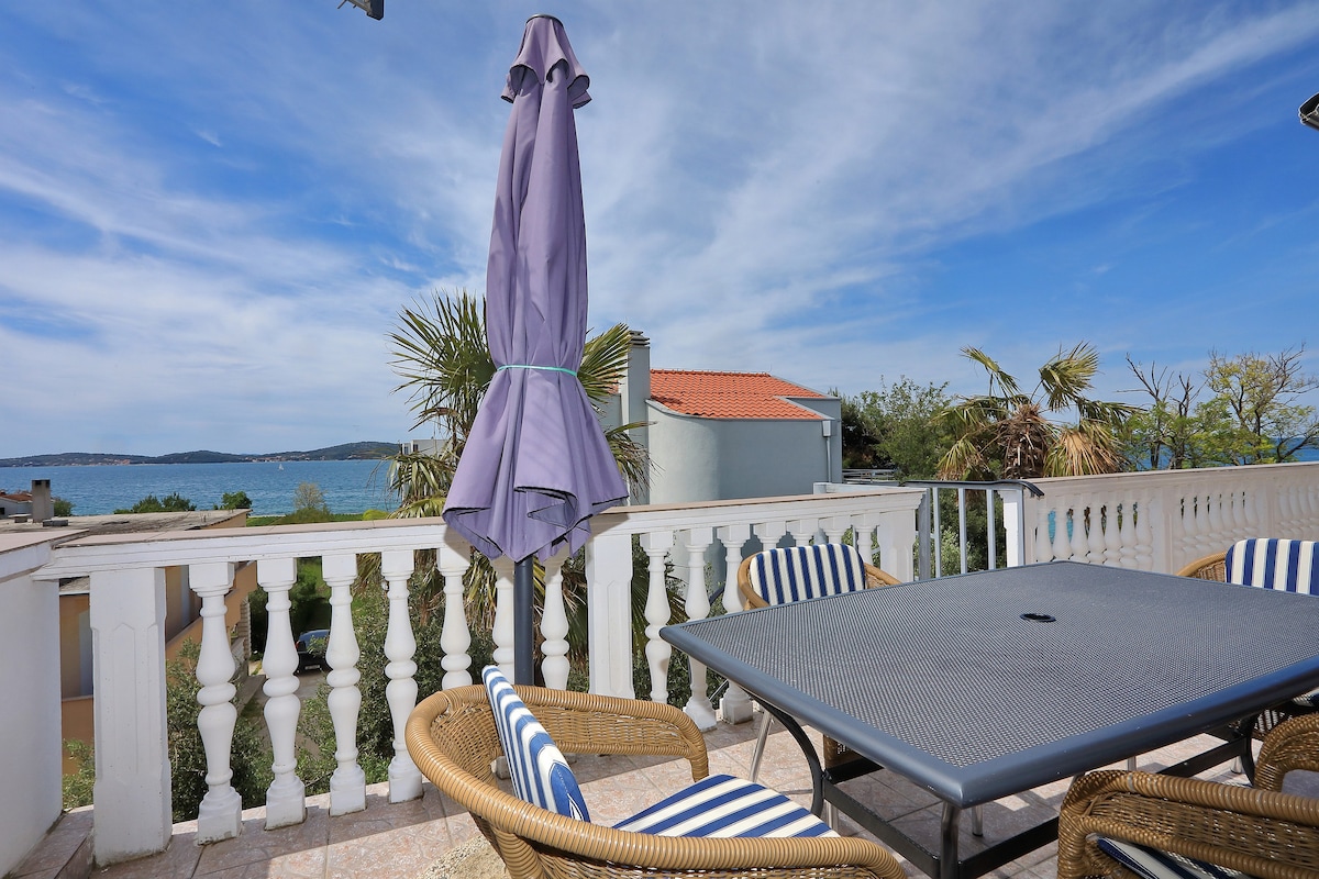 A terrace is presented with a dining table surrounded by striped chairs. A purple umbrella offers shade, and views of the sea and nearby islands are visible in the background. Lush greenery adds a touch of tranquility to the scenery.