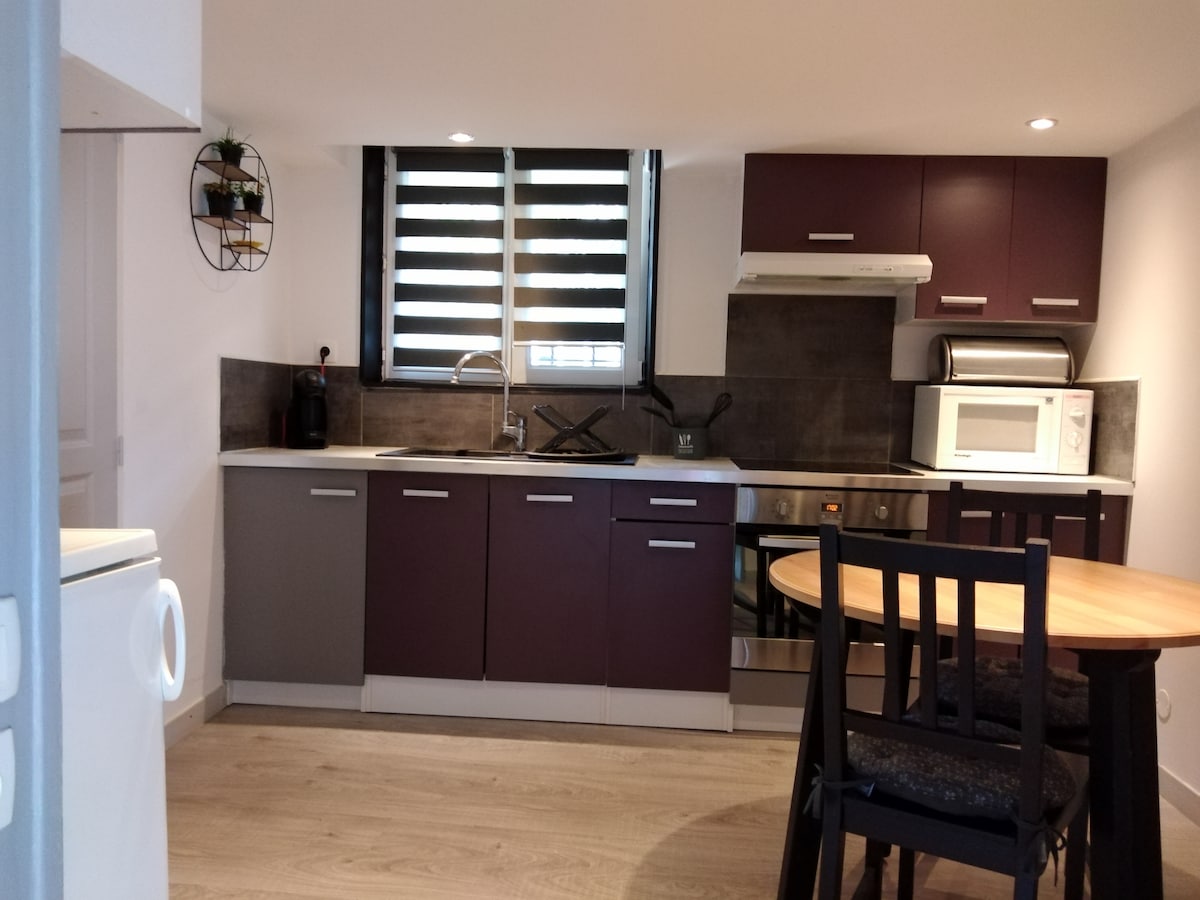 A modern kitchen is equipped with dark cabinets and light countertops. Essential appliances include a microwave and stovetop. A dining table with four chairs is positioned nearby, and light enters through a window with adjustable slats, creating a functional cooking space.