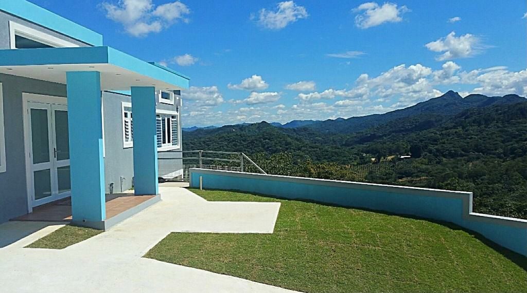 A modern house with a blue and gray exterior is set against a backdrop of lush mountains and a clear sky. A well-manicured lawn extends in front, providing ample outdoor space. The design includes large windows, allowing natural light to fill the interior.