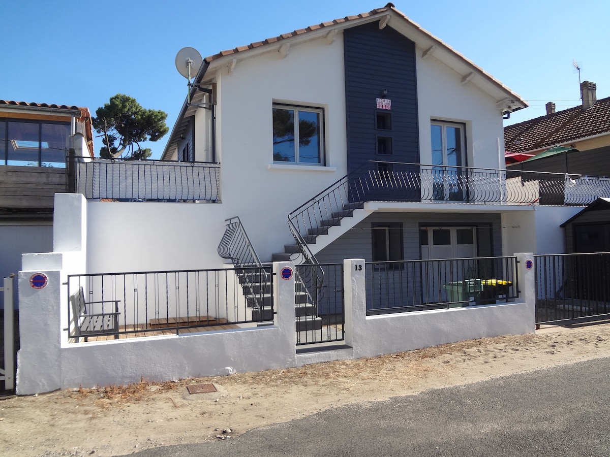 A two-story house features a white and dark blue exterior. A decorative railing surrounds the entrance and a staircase leads to the upper level. A satellite dish is mounted on the side. The surrounding area includes a small outdoor seating arrangement.