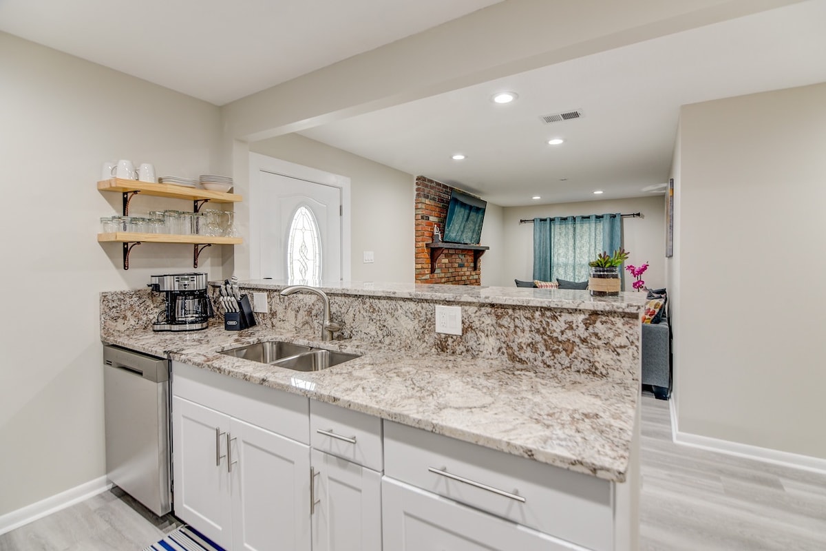 A modern kitchen features polished countertops with dual sinks, stainless steel appliances, and open shelving displaying glassware. A doorway leads to a welcoming living area, which is visible in the background, accentuated by a brick feature wall and light-colored furnishings.