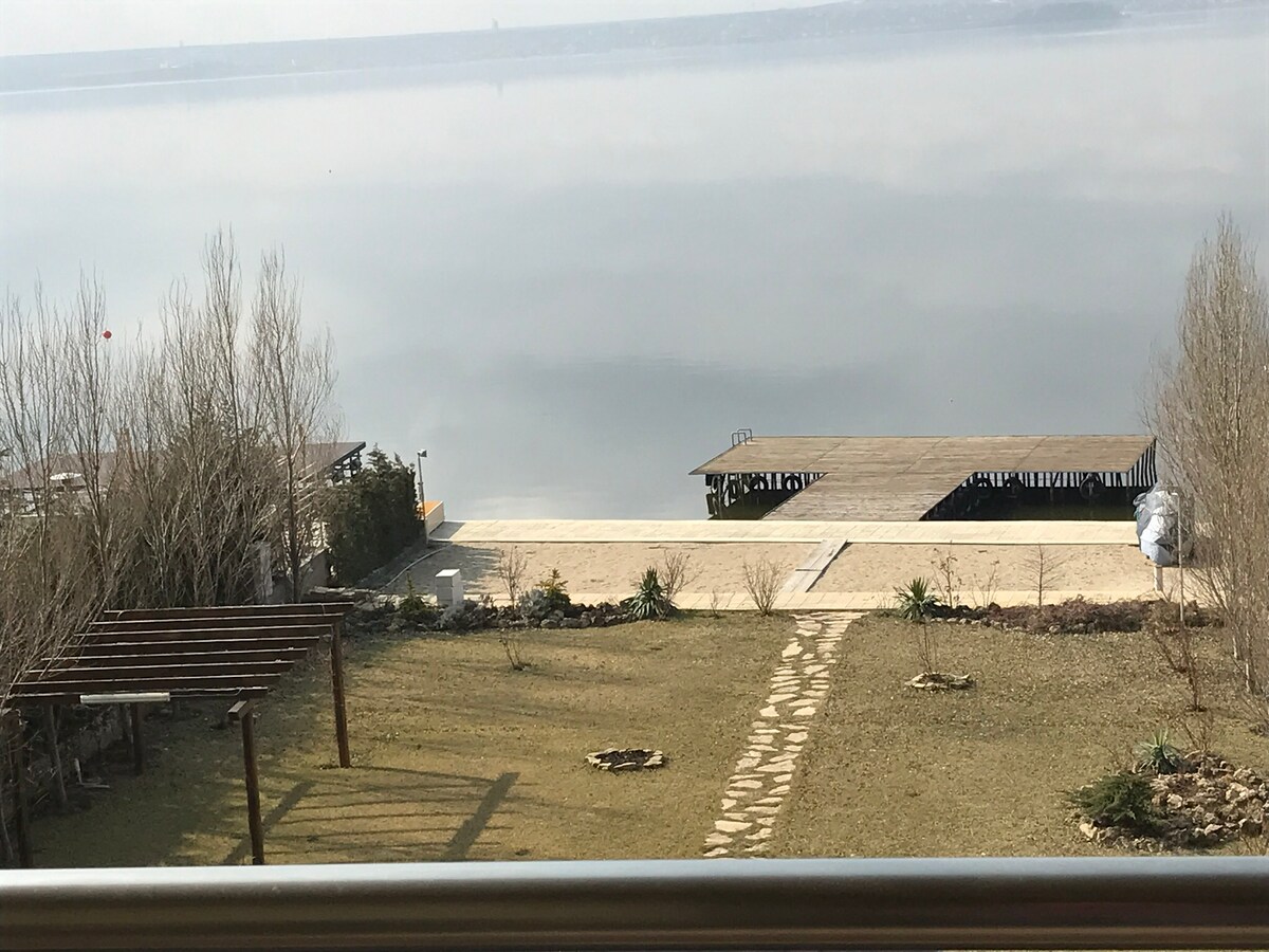 A serene outdoor space is visible, featuring a well-maintained lawn with stone pathways. A wooden deck extends towards a calm water body, reflecting the sky. Several bare trees and shrubs provide a natural border to the area, enhancing the tranquil ambiance.