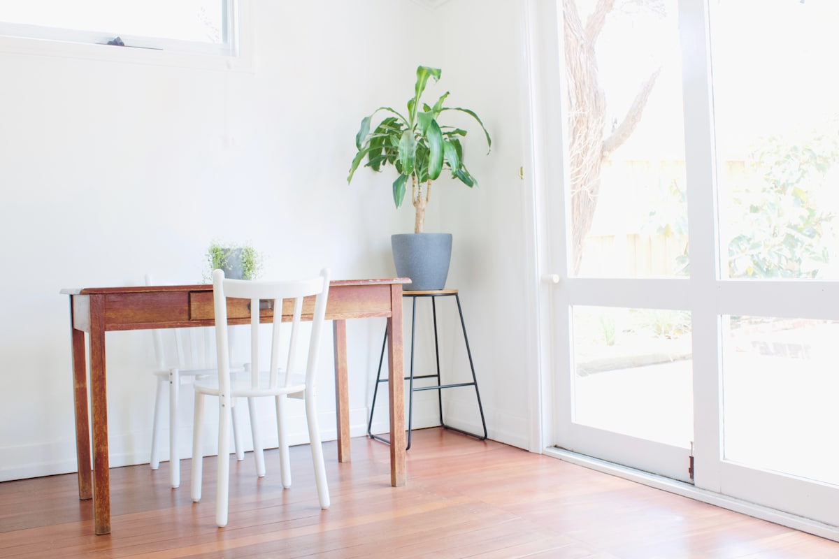 A dining area is presented with a wooden table and three chairs, accompanied by a potted plant on a stand. Natural light filters through large glass doors, illuminating the warm timber floor and offering a view of the outdoor garden.