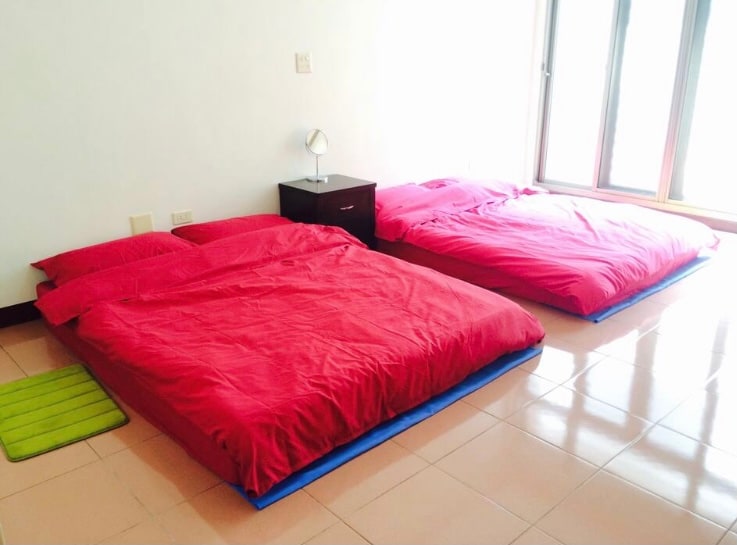 A spacious sleeping area contains two beds dressed in bright red bedding, positioned on a smooth tiled floor. A small nightstand stands beside one bed, and a green rug is placed nearby. Large windows allow natural light to fill the room.