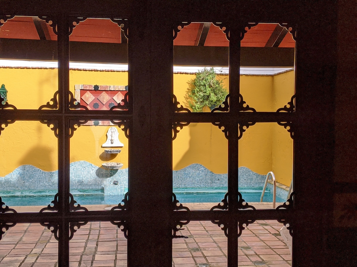 A view through ornate wooden doors reveals a tranquil courtyard featuring a swimming pool. The yellow walls create a warm ambiance, while a decorative fountain adds a serene element. Subtle greenery is visible in the background, enhancing the peaceful setting.