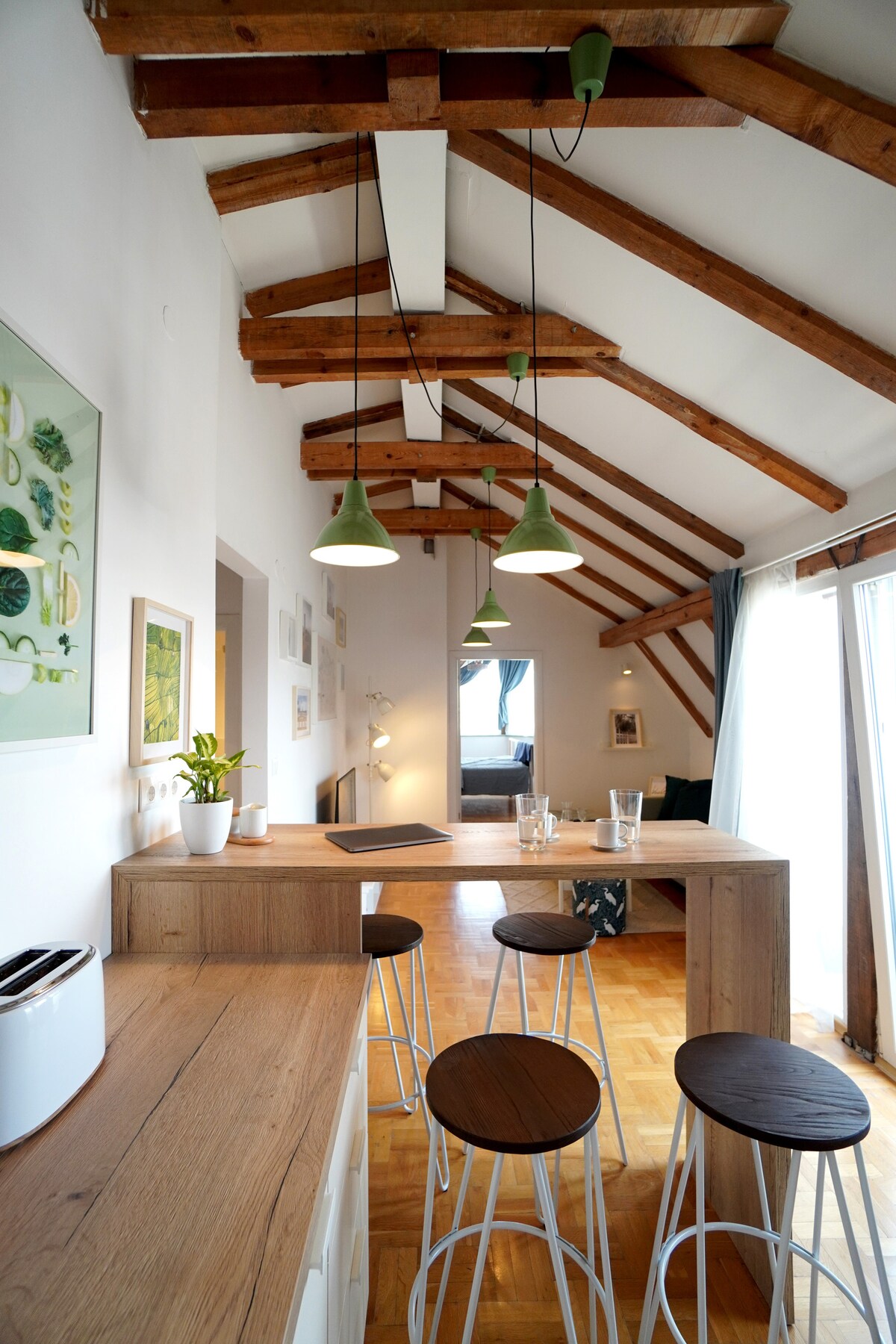 A spacious interior features exposed wooden beams and pendant lights in green hues. A light wood counter extends into a seating area with three minimalist stools. A bright window offers natural light, while decorative artwork and a plant add touches of character to the space.