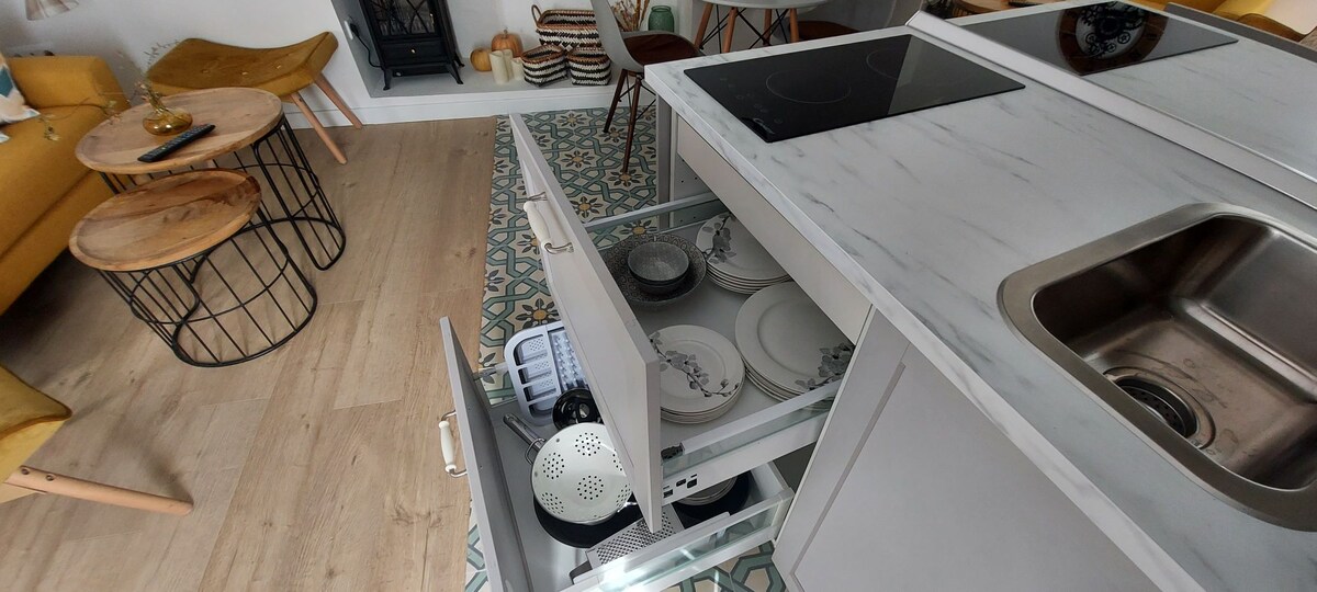 A modern kitchen space is highlighted, featuring a marble countertop with a cooktop and a stainless steel sink. An open drawer reveals plates and kitchen utensils. The floor displays a colorful patterned tile design, complemented by wooden tables and seating in the background.