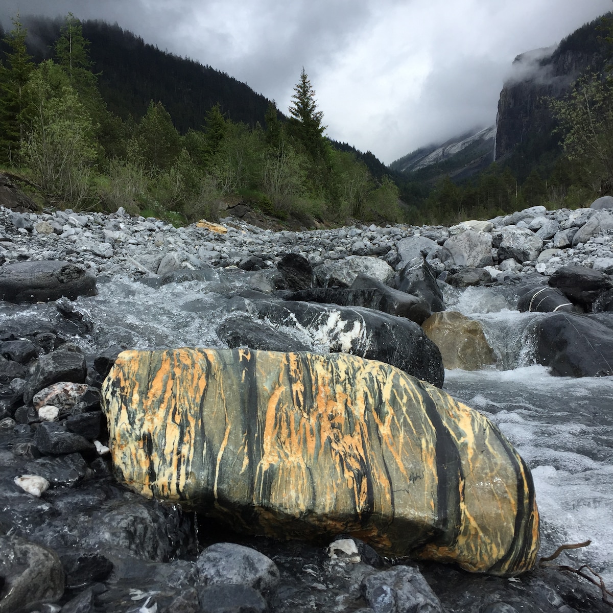 A large, striped rock with orange and yellow hues is positioned in a flowing river, surrounded by smooth pebbles and gravel. Lush green trees and mountainous terrain can be seen in the background, partially covered by clouds, enhancing the natural landscape.