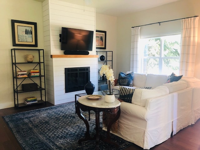 A cozy living room features a white sofa adorned with decorative pillows, positioned near a wall-mounted television. A fireplace is integrated into the shiplap surround, while a small round table with a floral arrangement sits on a patterned rug, creating an inviting atmosphere.