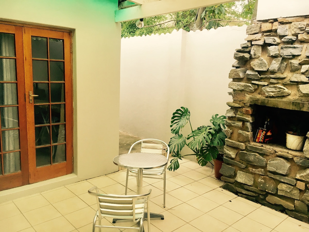 An outdoor seating area is visible, featuring a small round table accompanied by two metal chairs. A stone fireplace is integrated into the wall, with greenery placed nearby. Natural light enters through wooden doors, highlighting the tiled floor and neutral-colored walls.