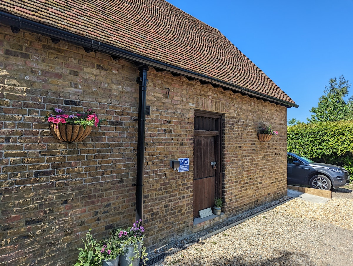 The exterior of Myrtle Stables is visible, featuring rustic brick walls and a wooden door with a traditional design. Flower baskets are mounted beside the entrance, and a parking area is adjacent, accommodating a vehicle. Clear blue skies enhance the inviting appearance.