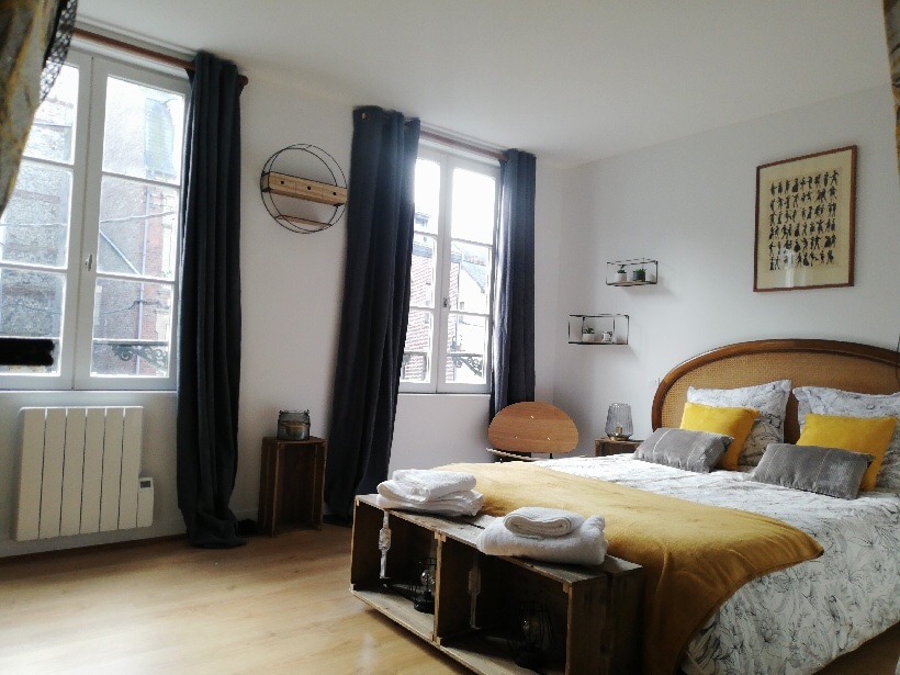 A bright bedroom features a spacious bed with a textured gray and white duvet, complemented by yellow cushions. Two large windows allow natural light to flood the room, while dark curtains frame the view. Wooden elements, including a bedside table and a unique wall shelf, enhance the decor.