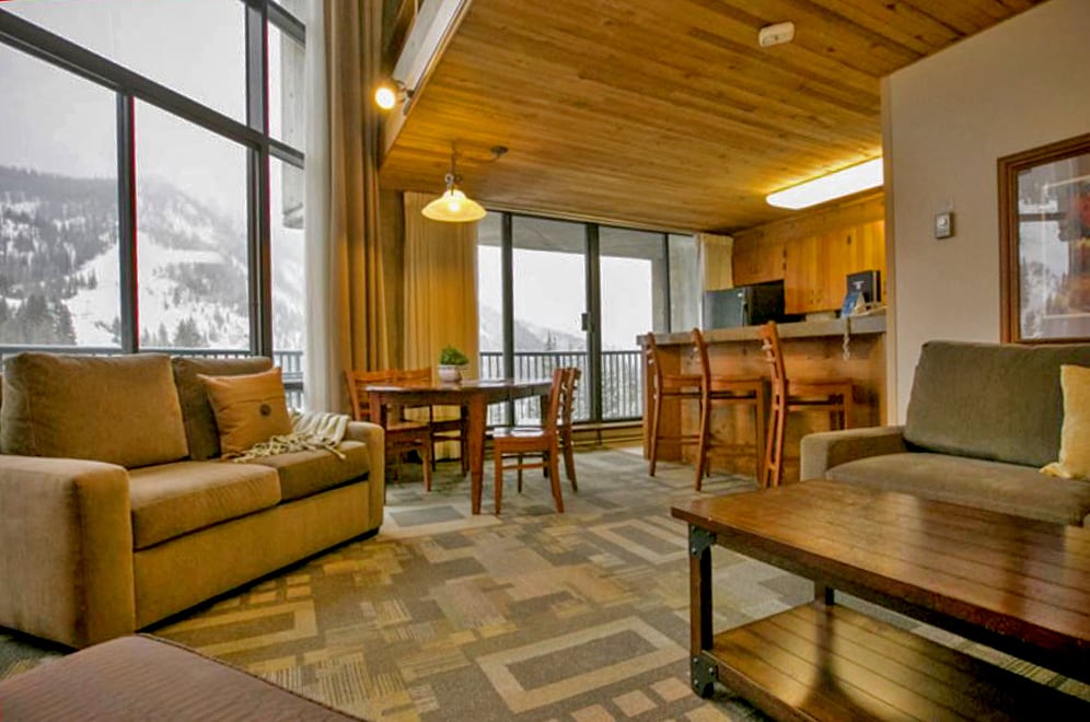 A spacious living area features a cozy sofa and armchair, complemented by a wooden coffee table. Large windows invite natural light and offer views of snow-covered slopes. A dining table with chairs and a kitchen bar are visible in the background.