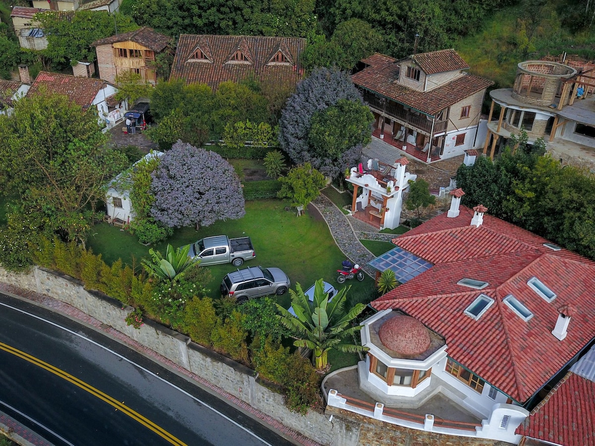 An aerial view reveals a scenic hostal surrounded by lush greenery and colorful trees. Various buildings with terracotta roofs are positioned around a manicured lawn, with several vehicles parked on the property. A winding road passes through the landscape, enhancing the natural setting.