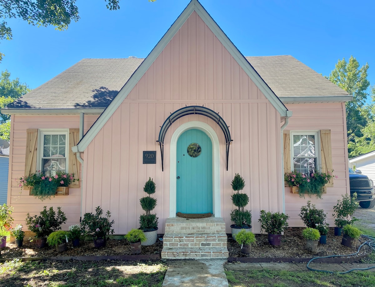 A charming pink cottage is featured, showcasing a light blue door and decorative plant boxes beneath the windows. Freshly manicured bushes and flowers border the entrance, with a brick pathway leading to the steps. The home is set against a clear blue sky.
