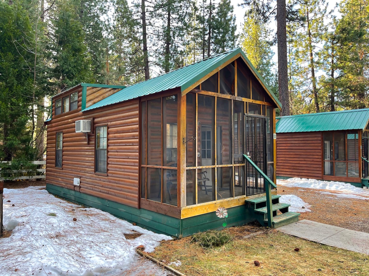 A charming cabin features a yellow door and a screened porch with a bench. Tall trees frame the exterior, while remnants of snow are visible on the ground. A similar cabin is positioned nearby, showcasing the rustic atmosphere of the area.