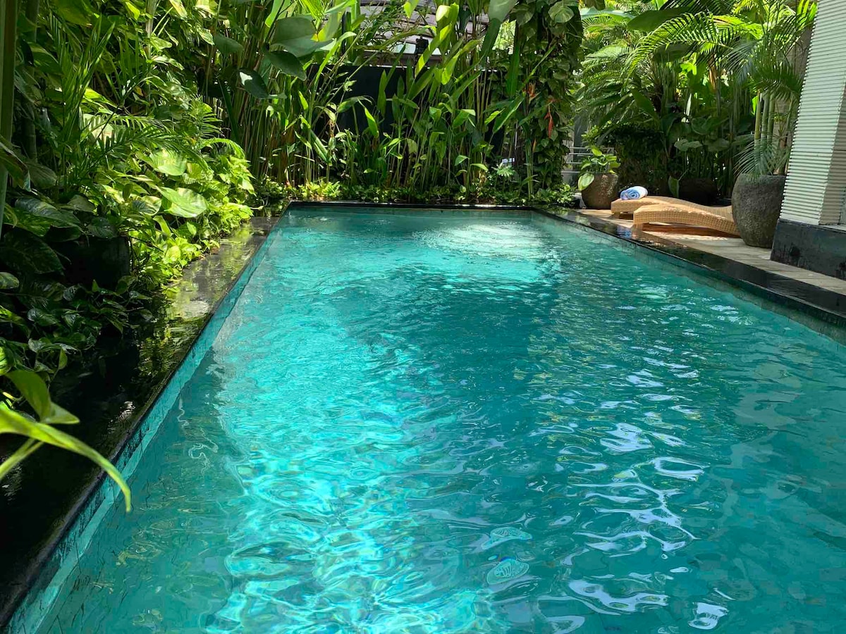 A private swimming pool surrounded by lush tropical greenery offers a serene atmosphere. The clear water reflects the sunlight, creating a calming blue hue. Various plants frame the pool, enhancing the connection to nature.