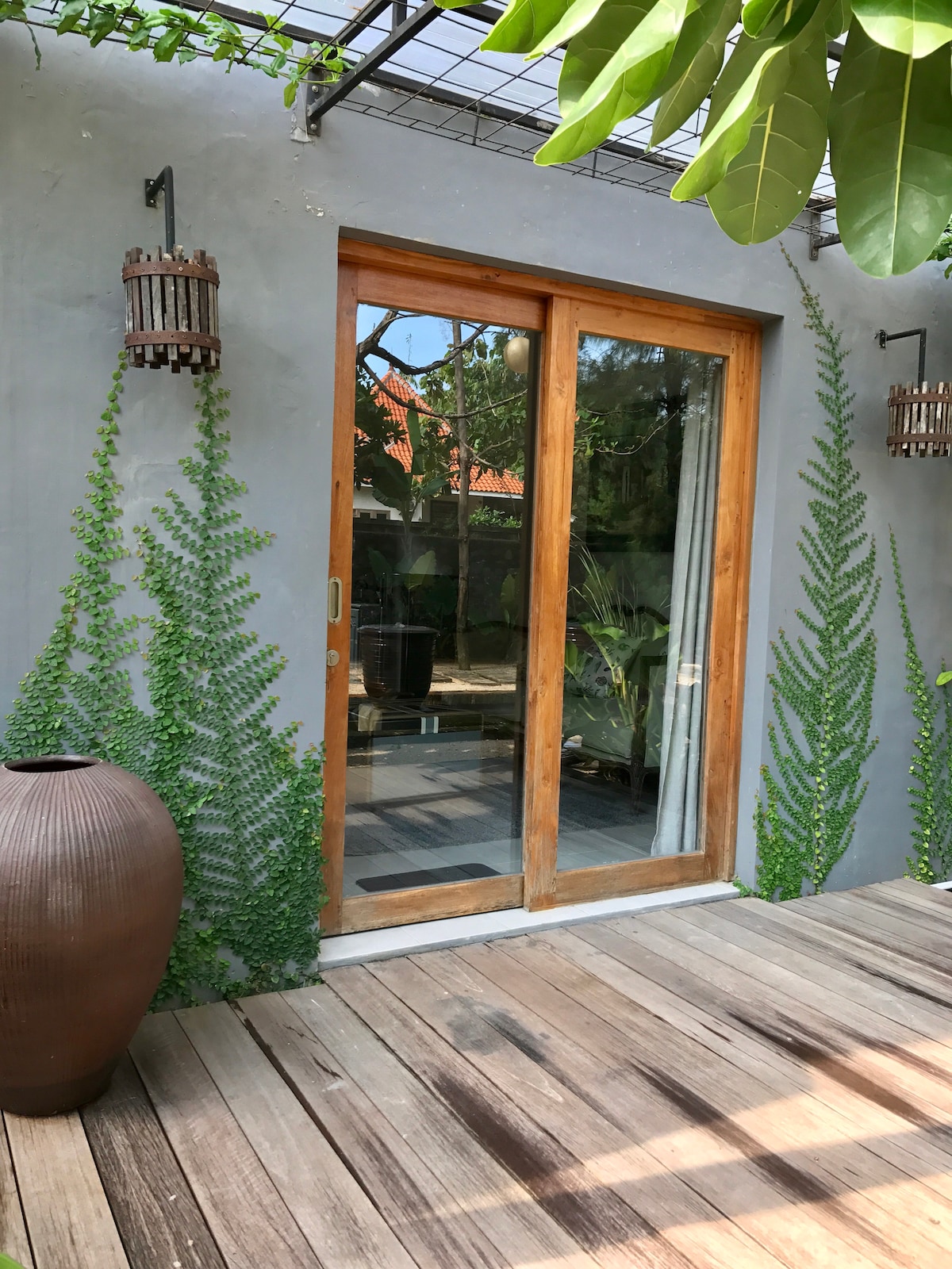 A wooden deck terrace is shown, leading to large glass doors framed by lush greenery. Two hanging lanterns are mounted on the gray walls beside the entrance, and a large, round planter sits on the deck, contributing to a natural ambiance.