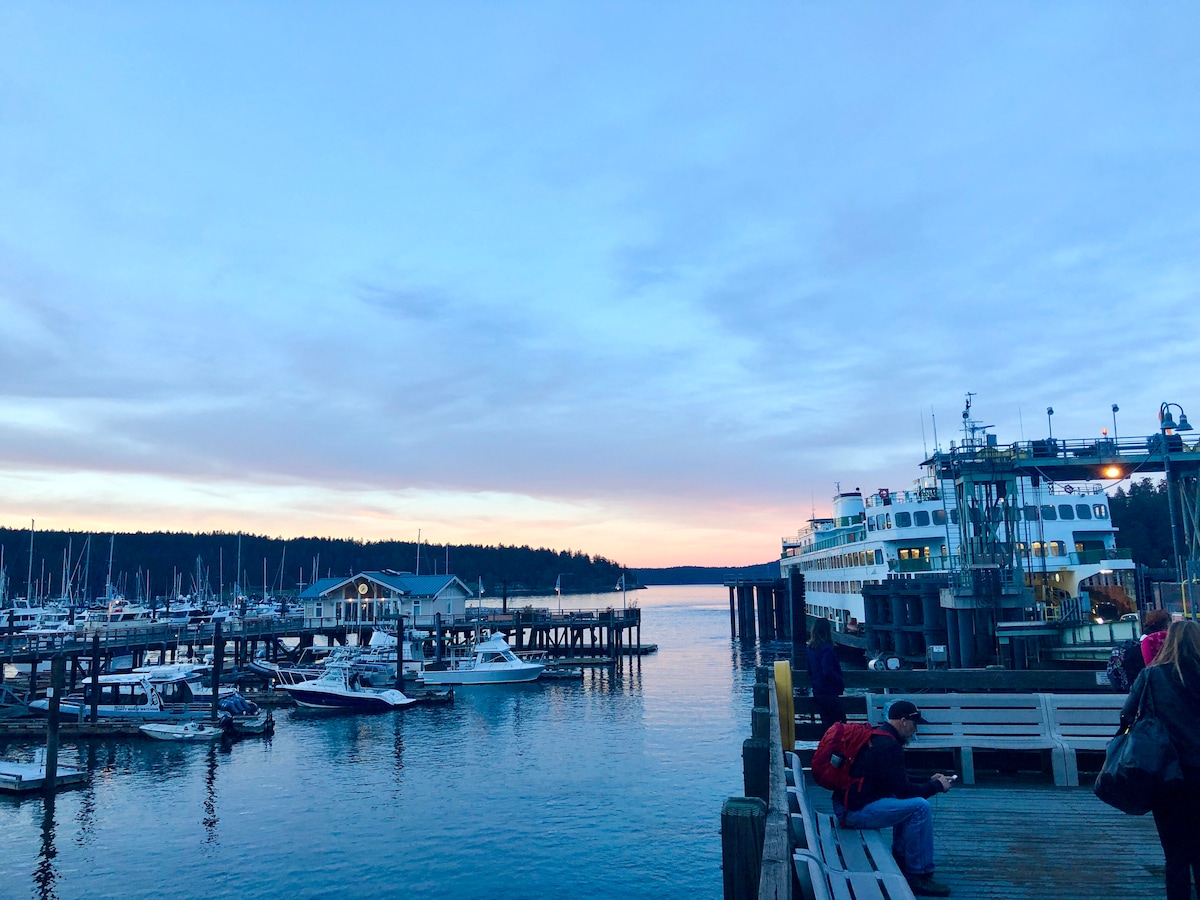 A serene waterfront scene is depicted at twilight, featuring a marina lined with boats. The calm water reflects soft hues of blue and pink as twilight approaches. A ferry is docked nearby, with a wooden boardwalk leading to the pier, inviting leisurely strolls.