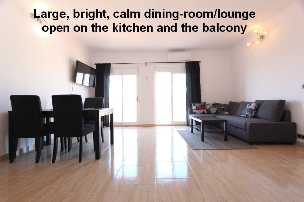 A spacious dining room and lounge area features light-colored tile flooring and is filled with natural light. A black dining table with chairs is visible, alongside a comfortable sofa. Large sliding doors open to a balcony, complementing the open layout connecting to the kitchen.
