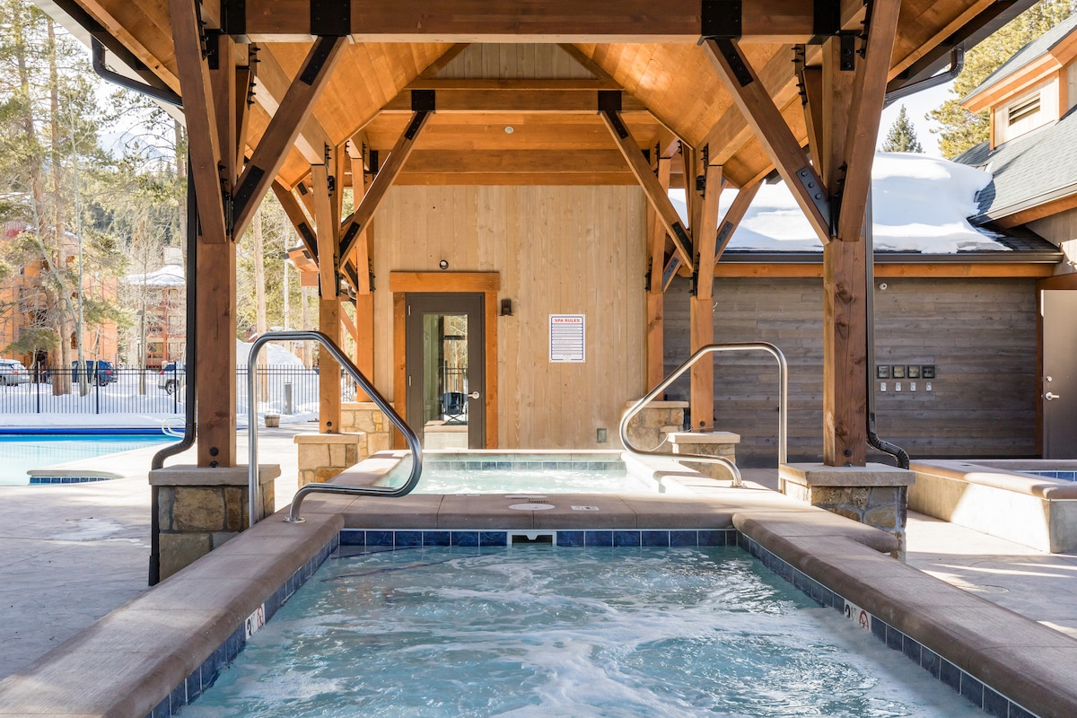A spacious hot tub is situated under a wooden pergola, showcasing a warm, bubbling surface. Nearby, the entrance to the community area is visible, along with a swimming pool in the background, surrounded by a wooden deck and trees.