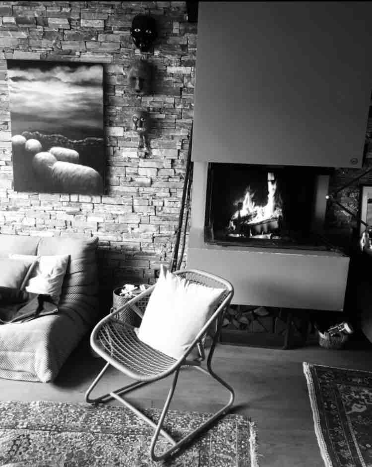 A cozy living room is highlighted by a modern fireplace, with a fire softly flickering. Textured stone walls serve as a backdrop to a colorful wall art piece and a comfortable chair adorned with a decorative pillow. A patterned rug adds warmth to the wooden floor.