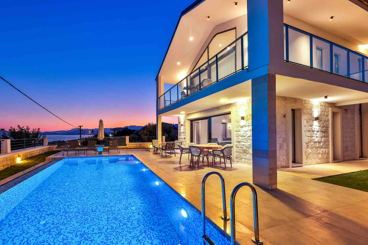 An exterior view of the villa showcases a modern two-story structure with large glass doors. A brightly lit outdoor pool reflects the evening sky, while a dining area with table and chairs is positioned nearby. Balconies extend from the upper floor, overlooking the landscape.