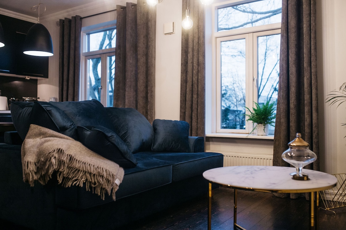 A dark blue sofa is positioned near large, bright windows adorned with sheer curtains. A cozy throw blanket is draped over the armrest. A marble-top coffee table with a decorative glass piece sits in front, enhancing the modern living space's elegance.