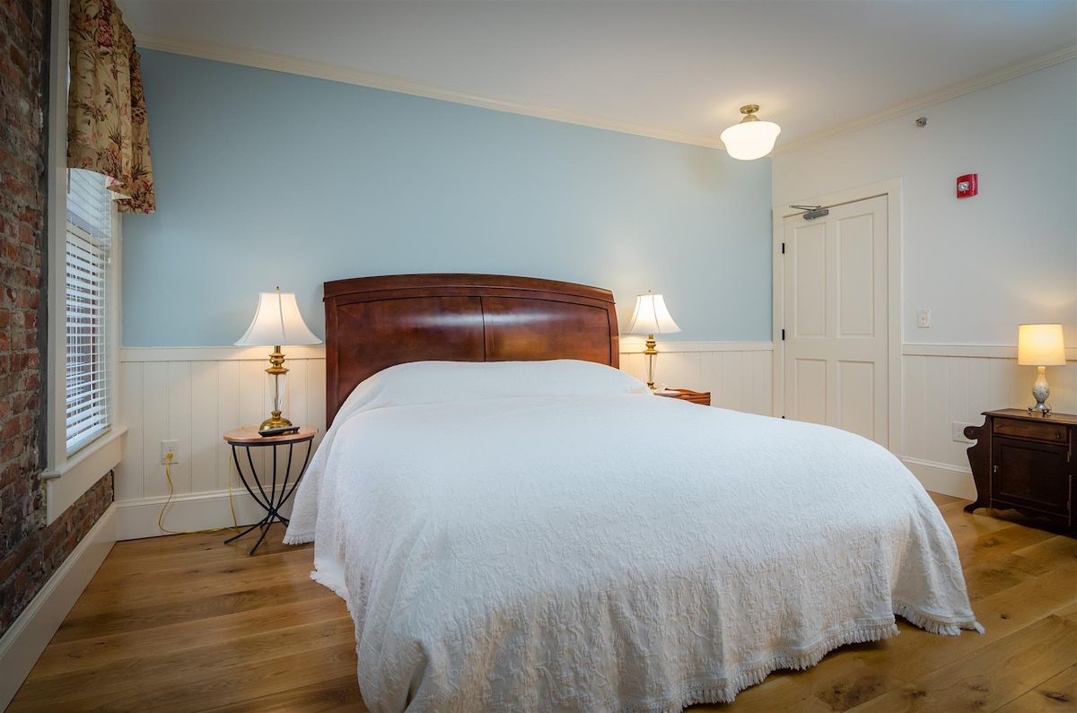 The room features a King-sized bed with a white quilted coverlet, accompanied by two bedside tables with lamps. A light blue wall provides a calming backdrop, while natural light filters in through a window, enhancing the warm wooden flooring.