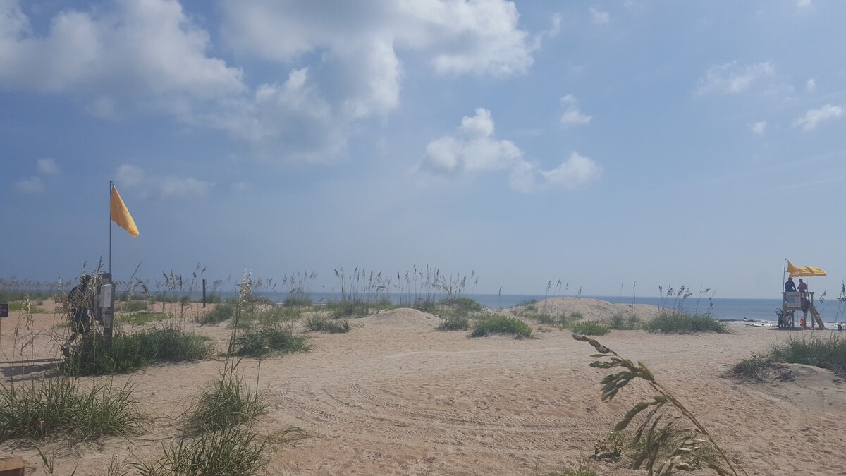 A sandy beach area is framed by tall grasses and gentle dunes under a bright sky. Yellow flags wave in the breeze near a lifeguard tower, indicating safety measures. The ocean is visible in the distance, with soft waves lapping at the shore.