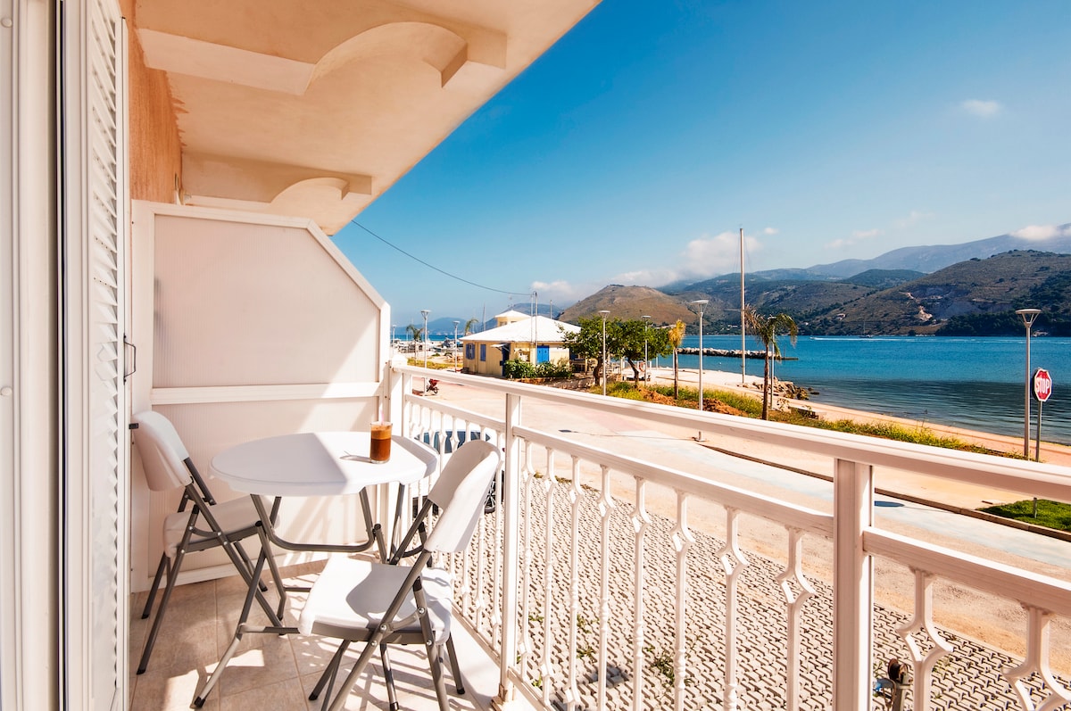 A balcony overlooking the sea features a small round table and two folding chairs. A scenic view includes the sparkling water, a sandy beach, and distant mountains under a clear blue sky. A pedestrian pathway and bicycle lane are visible along the waterfront.