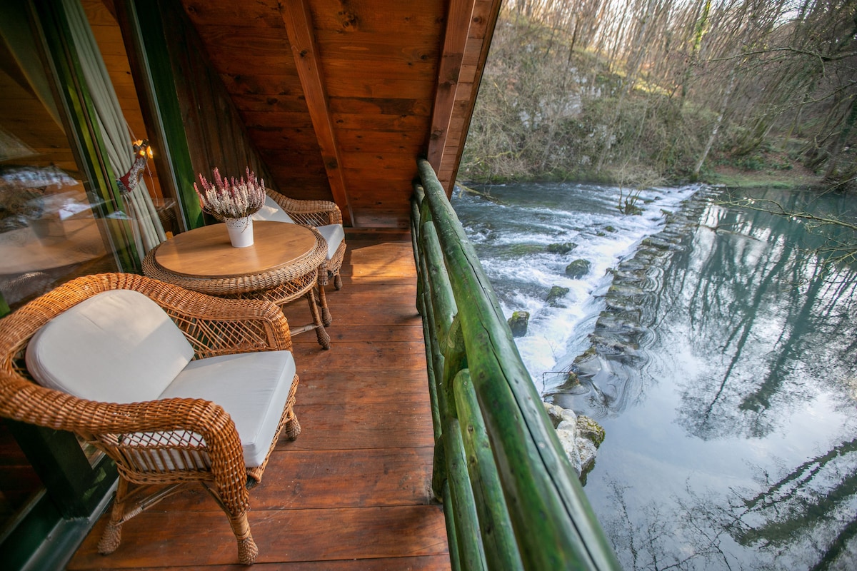 A cozy balcony is seen overlooking the river, featuring a round wooden table and two wicker chairs. The natural surroundings include flowing water and greenery, creating a serene atmosphere. Sunlight filters through the trees, enhancing the peaceful view.